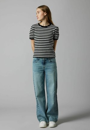 Svart- och vitrandig kortärmad stickad topp som bärs med ljusblå vida jeans. Vita sneakers synliga, mot en neutral bakgrund.