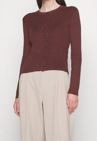 Geribbelde bordeauxrode cardigan met lange mouwen, ronde halslijn en bruine knopen aan de voorkant, gecombineerd met beige geplooide broek.