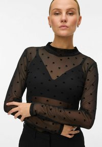 Zwarte mesh lange mouw top met een hoge hals, voorzien van een hartpatroon en een contrasterende effen zwarte onderlaag. Getextureerde stof en aansluitend ontwerp.