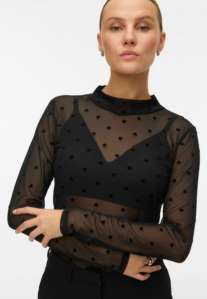 Zwarte mesh lange mouw top met een hoge hals, voorzien van een hartpatroon en een contrasterende effen zwarte onderlaag. Getextureerde stof en aansluitend ontwerp.