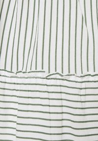 Valkoinen kangas, jossa on vuorottelevia vihreitä pystysuoria ja vaakasuoria raitoja, erotettuna kokoonpannaulutulla sauma, jossa on röyhelöreunus.