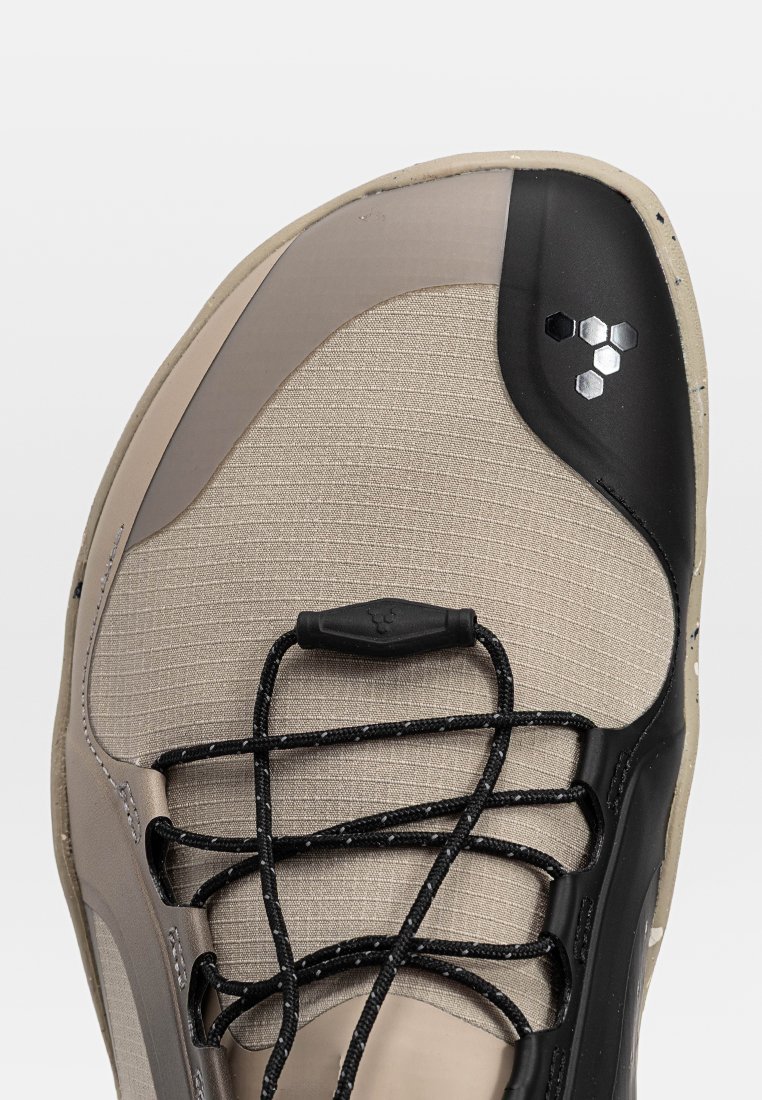 Zapato de escalada ligero con una parte superior beige texturizada, punta de goma negra y cordones negros para un ajuste seguro. Detalle del logo hexagonal presente.
