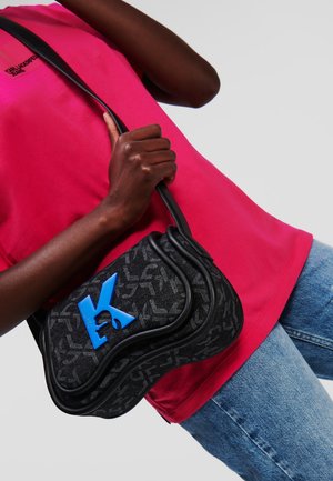 Borsa nera con motivo a fantasia e dettaglio blu a forma di 'K'; tessuto morbido, forma strutturata e una sottile tracolla nera, indossata sopra una maglietta rosa e jeans.