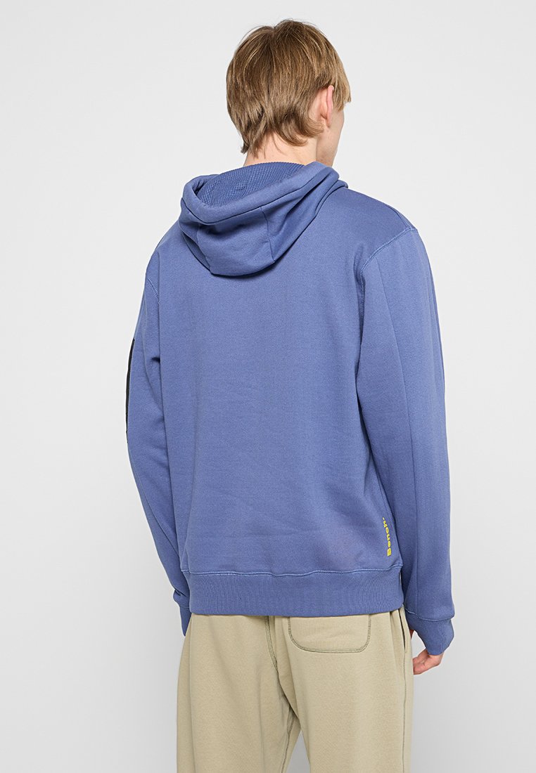 Sudadera de algodón azul con capucha ajustable, puños acanalados y un dobladillo con un pequeño acento de logo amarillo. Textura suave.