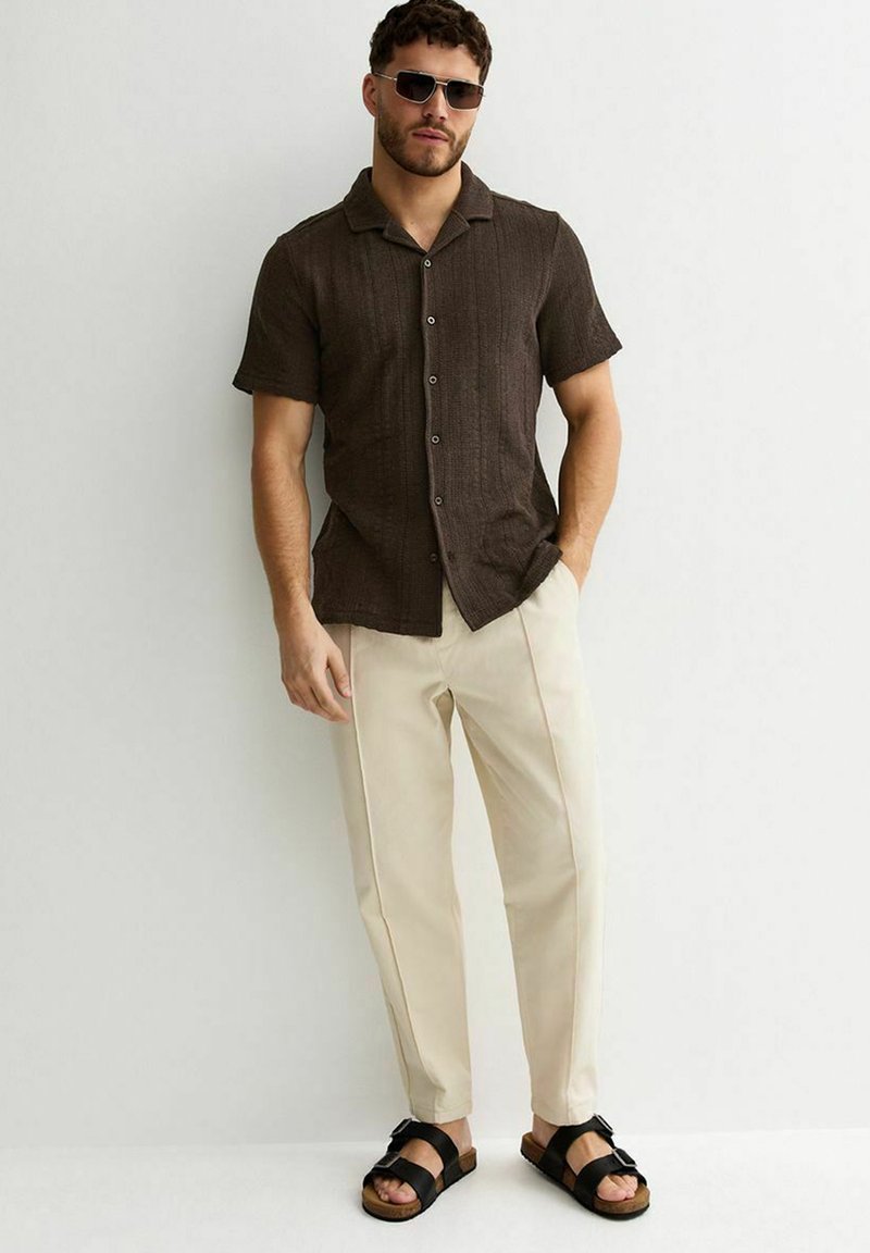 Uomo con camicia marrone a maniche corte con bottoni, pantaloni beige, sandali neri e occhiali da sole, in piedi contro un muro bianco.