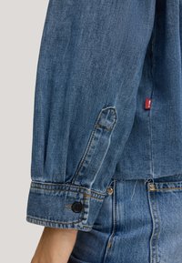 Blue denim jacket sleeve with a button closure and visible stitching. Features a red tag on the side. Smooth texture and tailored fit.