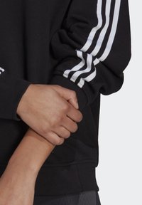 Svart tröja med vita ränder på ärmarna. Har mjukt tyg, lös passform och ribbad mudd. Händerna håller om ärmen.