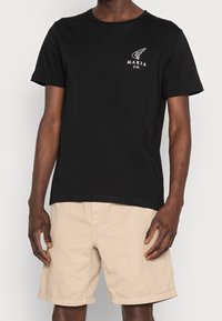 T-shirt en coton noir avec un logo imprimé blanc sur la poitrine gauche. Manches courtes et col rond. Pantalon beige.