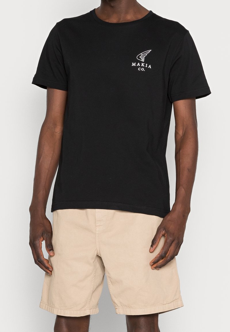 T-shirt en coton noir avec un logo imprimé blanc sur la poitrine gauche. Manches courtes et col rond. Pantalon beige.
