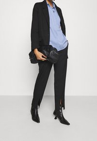Black tailored blazer over a light blue shirt, paired with black trousers featuring side slits, and pointed black ankle boots. Black clutch in hand.
