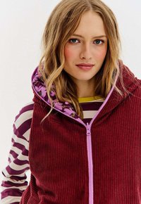 Młoda kobieta z falującymi blond włosami, ubrana w bordową kamizelkę z kordurowego materiału zapinaną na zamek, noszącą pod spodem bluzkę w fioletowo-białe paski.