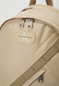 Zaino beige con un tessuto testurizzato, dotato di cerniere argentate, una toppa con logo e una tracolla a contrasto. Design compatto per un uso quotidiano.
