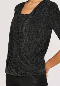 Haut texturé noir avec un col carré, des manches trois-quarts et un drapé à l'avant. Présente un léger éclat métallique.