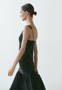 Donna con capelli raccolti in uno chignon, che indossa un top nero senza maniche con dettagli in pizzo e una gonna plissettata, che cammina di lato su uno sfondo chiaro uniforme.