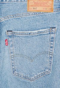 Tela de mezclilla azul clara con un bolsillo trasero con costuras amarillas y una etiqueta con el logo de Levi's que indica Lot 501, W32, L32.