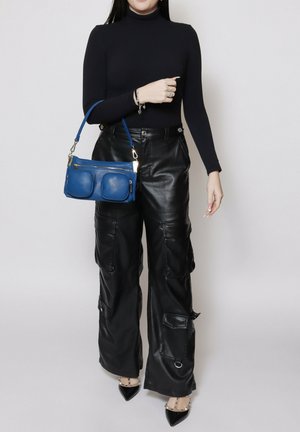 Donna che indossa un dolcevita nero e pantaloni cargo in pelle, tiene una borsa blu con doppia tasca frontale e indossa tacchi neri borchiati.