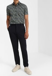 Camicia a maniche corte con motivo floreale in blu scuro e crema, abbinata a pantaloni su misura color navy e sneakers chiare con suola texturizzata.