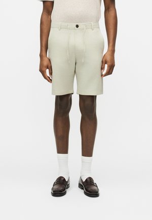 Homme portant un short beige à cordon, des chaussettes blanches et des mocassins marron foncé, debout devant un fond clair uni.