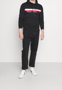 Sudadera negra con un diseño a rayas en rojo, blanco y azul, bolsillo delantero, combinada con jeans negros y zapatillas blancas. Corte casual, material de algodón.