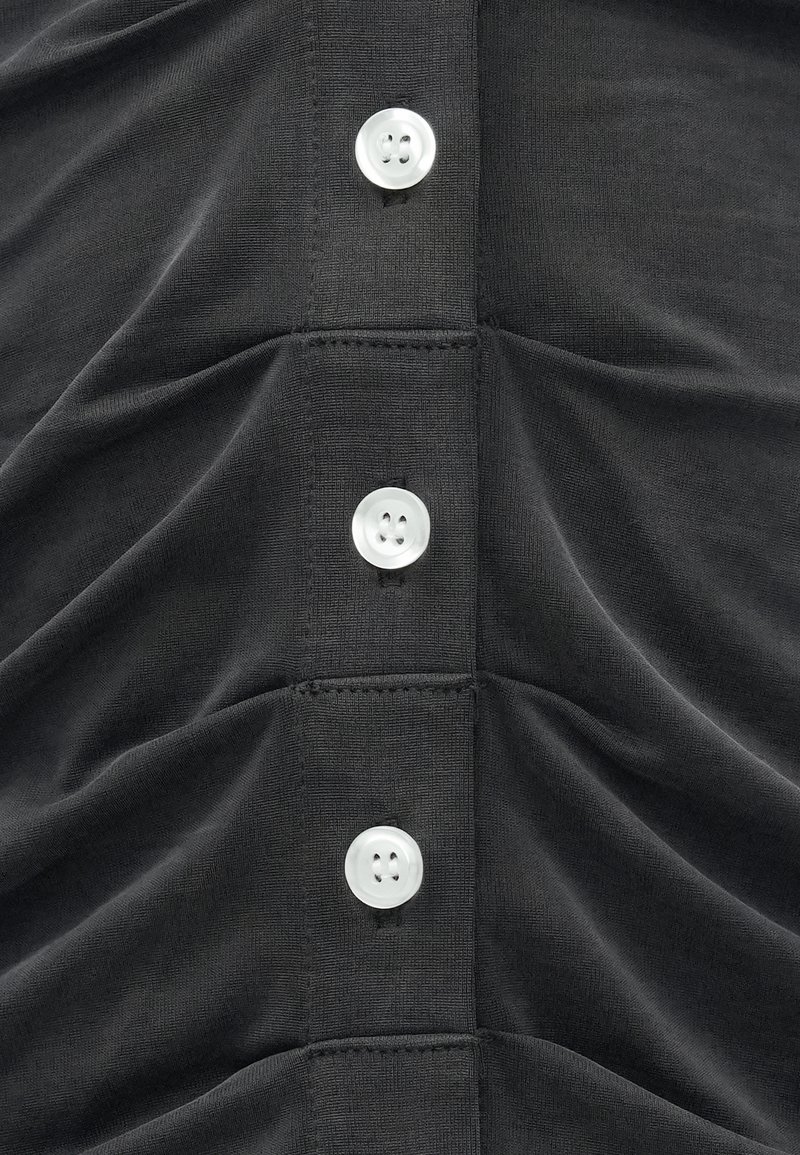 Sort, struktureret stoftop med en række af fire hvide cirkulære knapper og samlet detaljering langs den forreste søm.