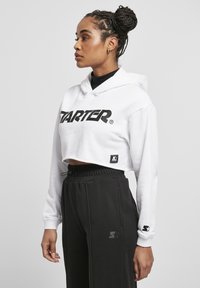 Hoodie bianco corto con un prominente logo nero "STARTER", abbinato a pantaloni della tuta neri. Include un patch con logo sul lato e una vita regolabile con coulisse.