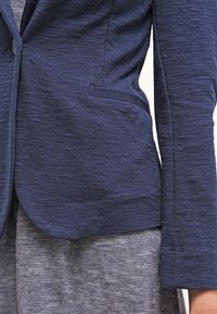 Granatowa marynarka z fakturowanej tkaniny, z pojedynczym zapięciem na guzik i boczną kieszenią, w zestawieniu z szaro-prążkowaną sukienką.