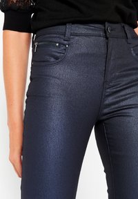 Tumma laivansininen kapea farkut, jotka on valmistettu kiiltävästä denimestä. Varusteena vetoketjullinen etutasku ja tavallinen farkkuommellus. Vähäisessä tekstuurissa, muotoa myötäilevä muotoilu.