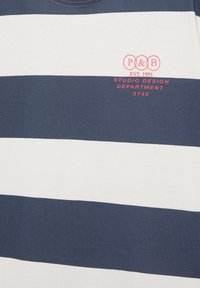 Marineblå og hvidt horisontalt stribet skjorte med rød tekst "P & B EST. 1991 Studio Design Department 3743" på brystet.