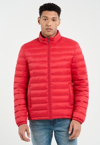 Rød puffjakke med høj krave, quiltet design og elastiske manchetter. Har en frontlynlås og sidelommer, parret med blå jeans.
