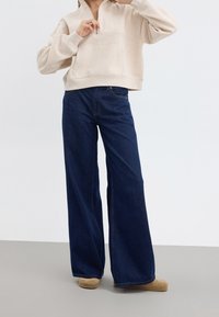 Beige kort hoodie med dragkedja i kragen och en framficka, tillsammans med mörkblå vida jeans och ljusbruna skor.