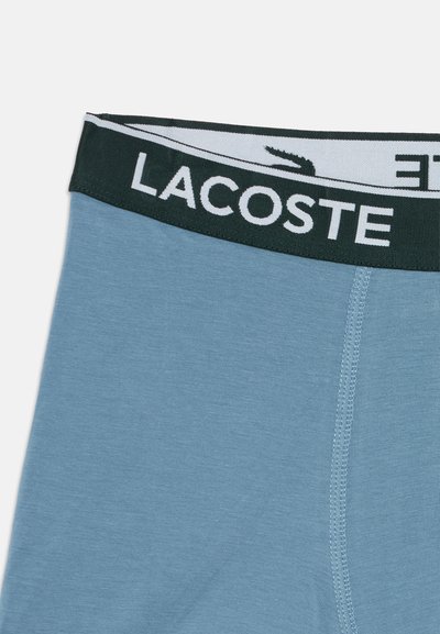 Tissu bleu clair avec une ceinture noire arborant le logo blanc Lacoste et une partie d'une icône de crocodile sur la bande.