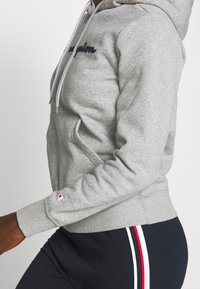 Person som bär en grå Champion-huvtröja med snoddar och svarta byxor med vita och röda ränder längs sidan.