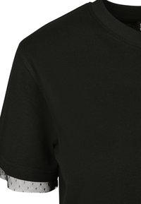 Haut noir à manches courtes avec col rond, orné d'un bord en maille noire transparente à pois qui dépasse sous les manches.