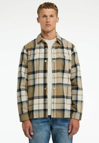 Hombre vestido con una camisa de cuadros en tonos beige, negro y blanco sobre una camiseta blanca y jeans azules, apoyado contra un fondo blanco liso.