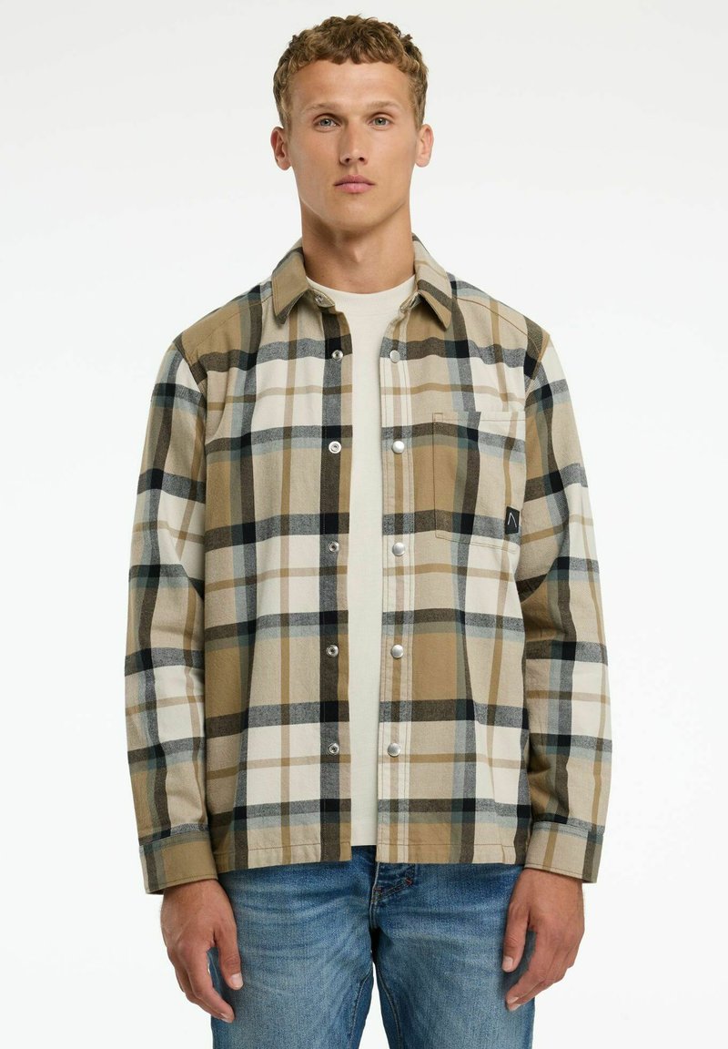 Hombre vestido con una camisa de cuadros en tonos beige, negro y blanco sobre una camiseta blanca y jeans azules, apoyado contra un fondo blanco liso.