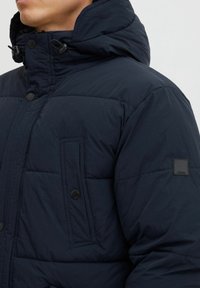 Chaqueta acolchada azul marino con cierre de cremallera y botón, que cuenta con capucha, textura acolchada y un bolsillo de parche en el lado izquierdo del pecho.