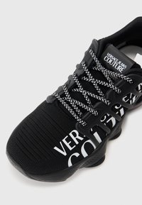 Μαύρο αθλητικό παπούτσι με πλέγμα στο πάνω μέρος, λευκό λογότυπο και κυματιστές μύτες. Σημαντικά χαρακτηριστικά περιλαμβάνουν χοντρές σόλες και κομψό σχεδιασμό.