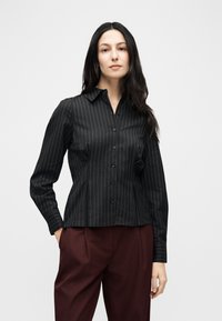 Γυναίκα με μακριά σκούρα μαλλιά, που φοράει μαύρο πουκάμισο με ρίγες και κόκκινο σκούρο παντελόνι, στέκεται με το ένα χέρι στην τσέπη μπροστά σε λευκό φόντο.