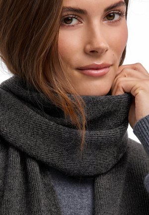 Young woman with light brown hair wearing a dark gray knitted scarf and sweater, gently holding the scarf near her chin.
