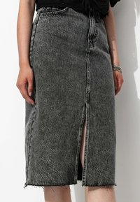 Jupe en denim noir taille haute avec une fente frontale, un ourlet effiloché et des détails de couture visibles. Le matériau semble robuste et texturé.