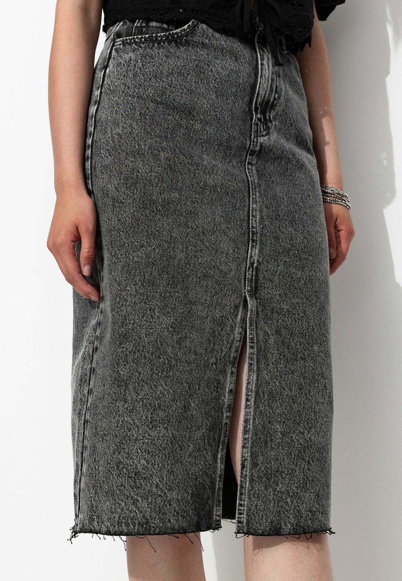 Jupe en denim noir taille haute avec une fente frontale, un ourlet effiloché et des détails de couture visibles. Le matériau semble robuste et texturé.