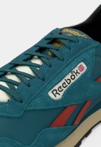 Ténis em camurça azul-petróleo com atacadores brancos, detalhes em vermelho e um logótipo da Reebok. Apresenta uma lingueta acolchoada e material texturizado para aderência.