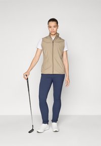 Beige sleeveless vest with a front zipper, paired with a white t-shirt and navy blue pants. White sneakers and a golf club are visible.