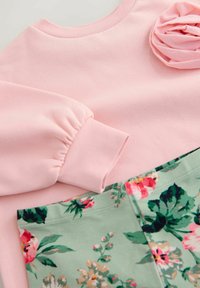 Roza sweatshirt z nabranimi rokavi in podrobnostjo iz cvetličnega blaga, v kombinaciji z zelenim materialom s cvetličnim vzorcem v odtenkih roza in zelene.