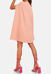 Mujer con un vestido corto color durazno estilo capa y sandalias rosas de tacón grueso con cordones, de pie frente a un fondo liso y claro.