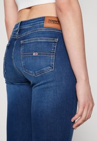Jeans de denim azul con una etiqueta de cuero que dice "TOMMY JEANS", que cuentan con dos bolsillos traseros y costuras en contraste en un diseño ajustado.