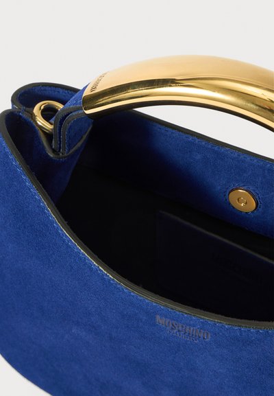Kék velúr kézitáska fekete díszítéssel, arany, ívelt fém fogantyúval, patentzárral és a "Moschino" felirattal az oldalán.