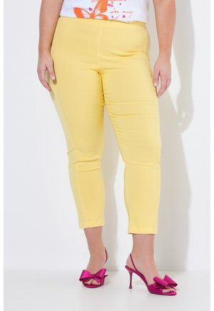 Person trägt gelbe Cropped-Hosen und knallpinke Sandalen mit hohen Absätzen und großen Schleifen, steht vor weißem Hintergrund.