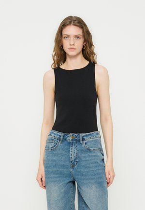 Ung kvinne med bølgete hår som har på seg gull øreringer i ringform, en ermeløs svart topp og blå jeans med høy midje mot en ensfarget bakgrunn.