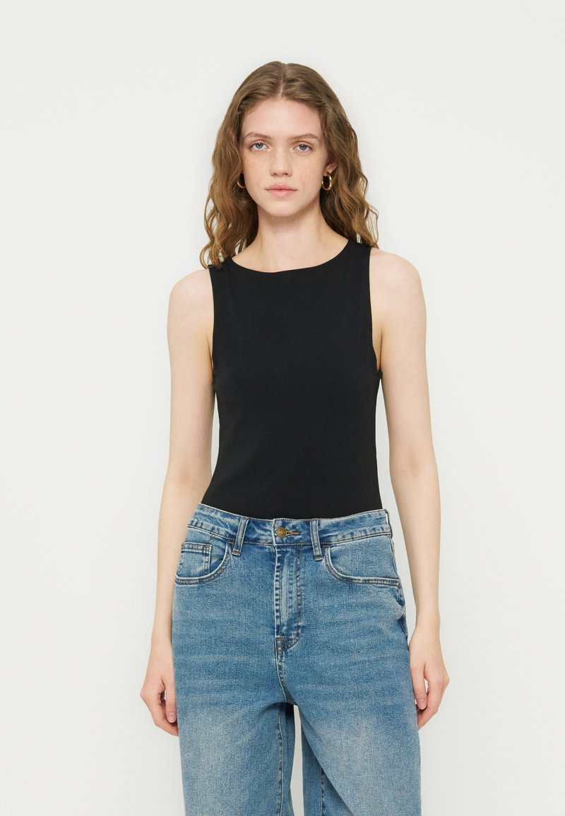 Νέα γυναίκα με κυματιστά μαλλιά που φορά χρυσούς κρίκους σκουλαρίκια, ένα μαύρο τοπ χωρίς μανίκια και ψηλόμεσα μπλε τζιν σε απλό φόντο.